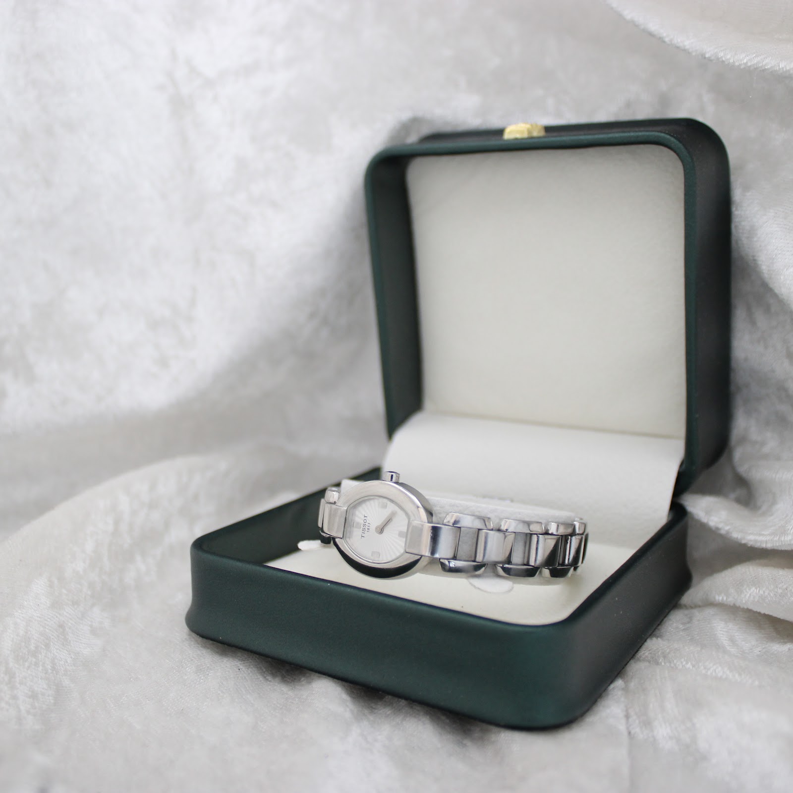 The Tissot G332 watch resting inside an open dark green presentation box with cream interior, set against a soft white textured background.