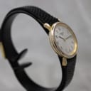 Angled view of the Tissot T852K watch from the left, showing the side profile of the gold-tone case and black leather strap on a watch stand.