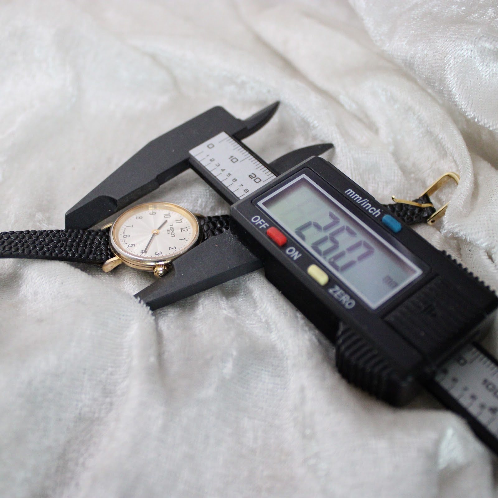 Overhead view of the Tissot T852K watch case being measured by a digital caliper, displaying a precise diameter of 26.0mm.