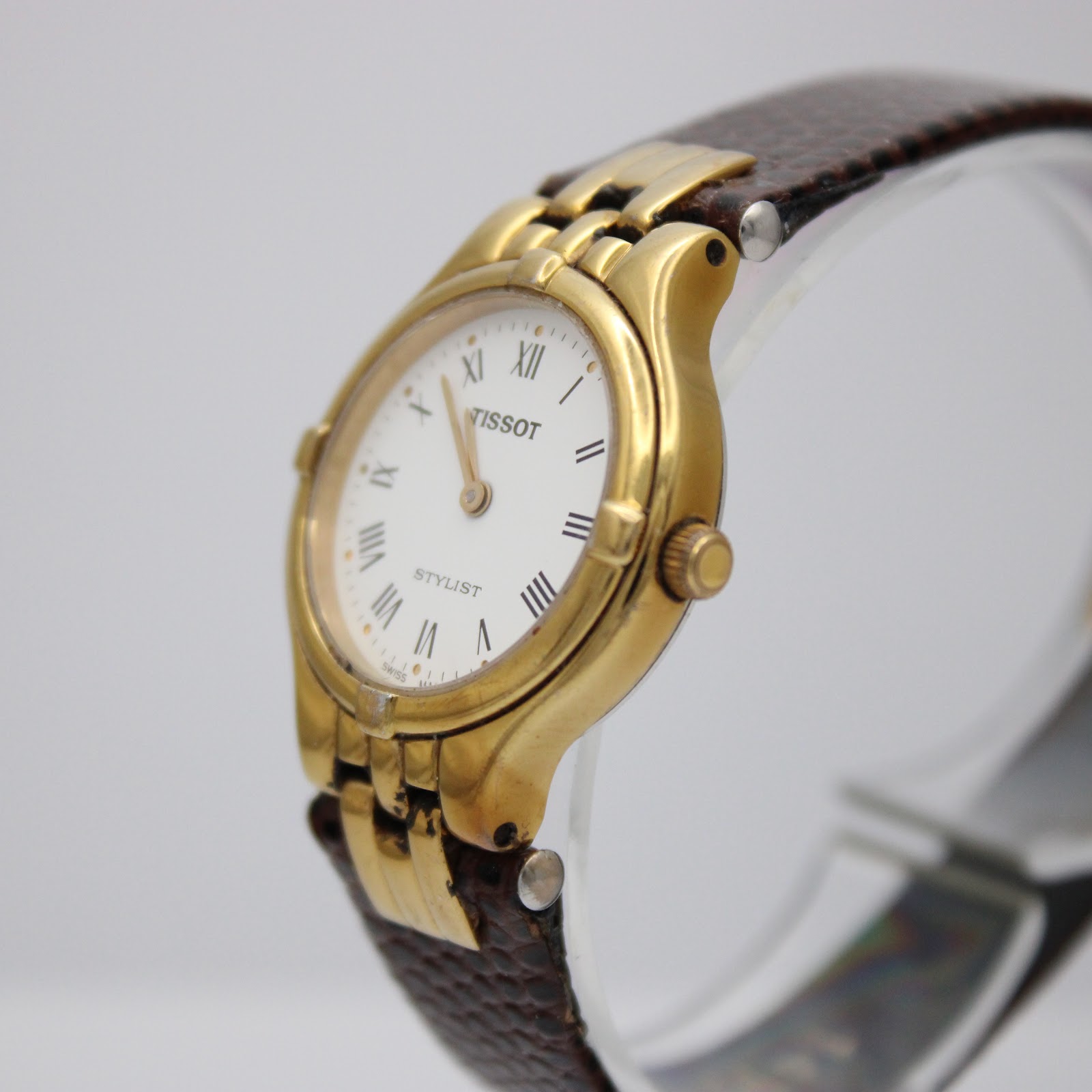 Angled side view of the Tissot Stylist watch, highlighting the gold-tone case and brown leather strap on a display stand.