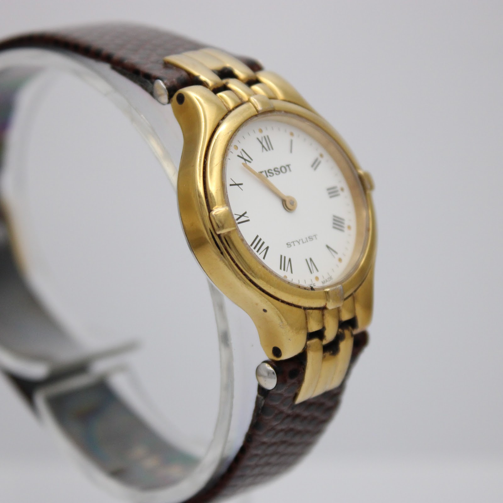Another angled side view of the Tissot Stylist watch, focusing on the curved gold-tone case and textured brown strap.
