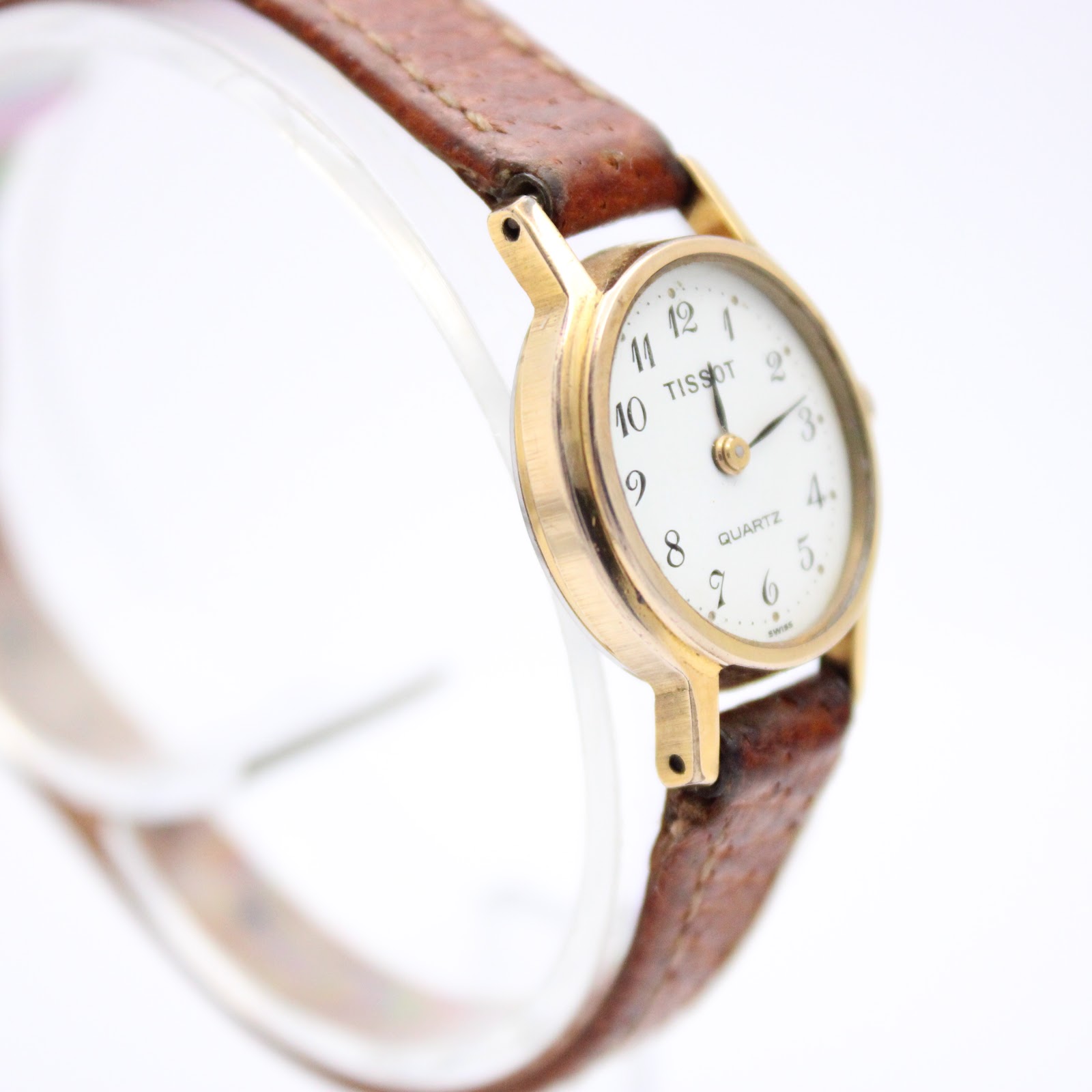 Close-up, slightly elevated angled view of the Tissot watch, focusing on the white dial, slender hour and minute hands, and the texture of the brown leather strap.