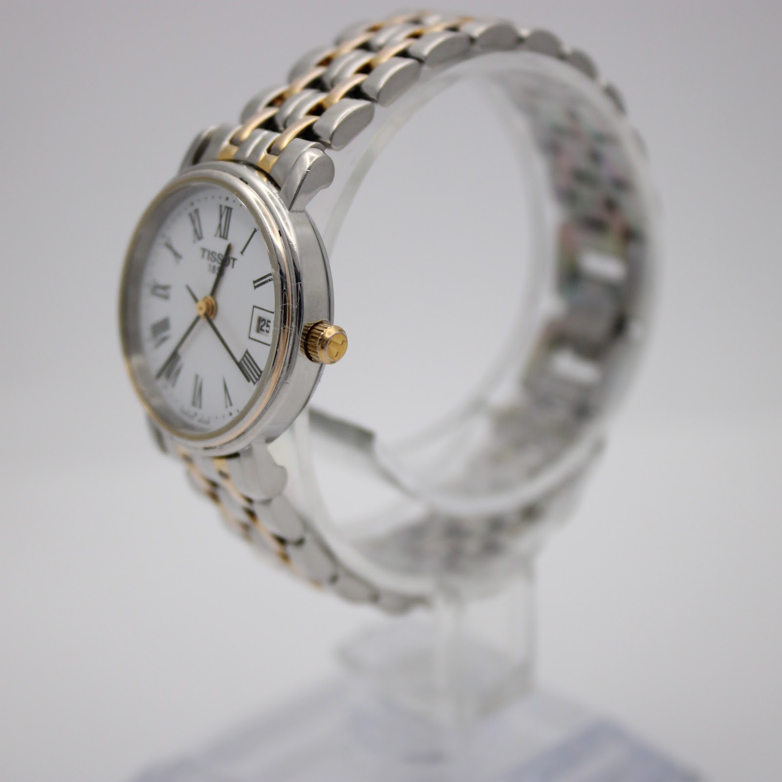 Angled side view of the Tissot 1853 watch on a clear display stand, highlighting the two-tone bracelet and gold-tone crown.