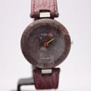 Front view of the Tissot Rock Watch R150 showing its dark red, natural stone dial and case, with yellow and red hands, on a watch stand.