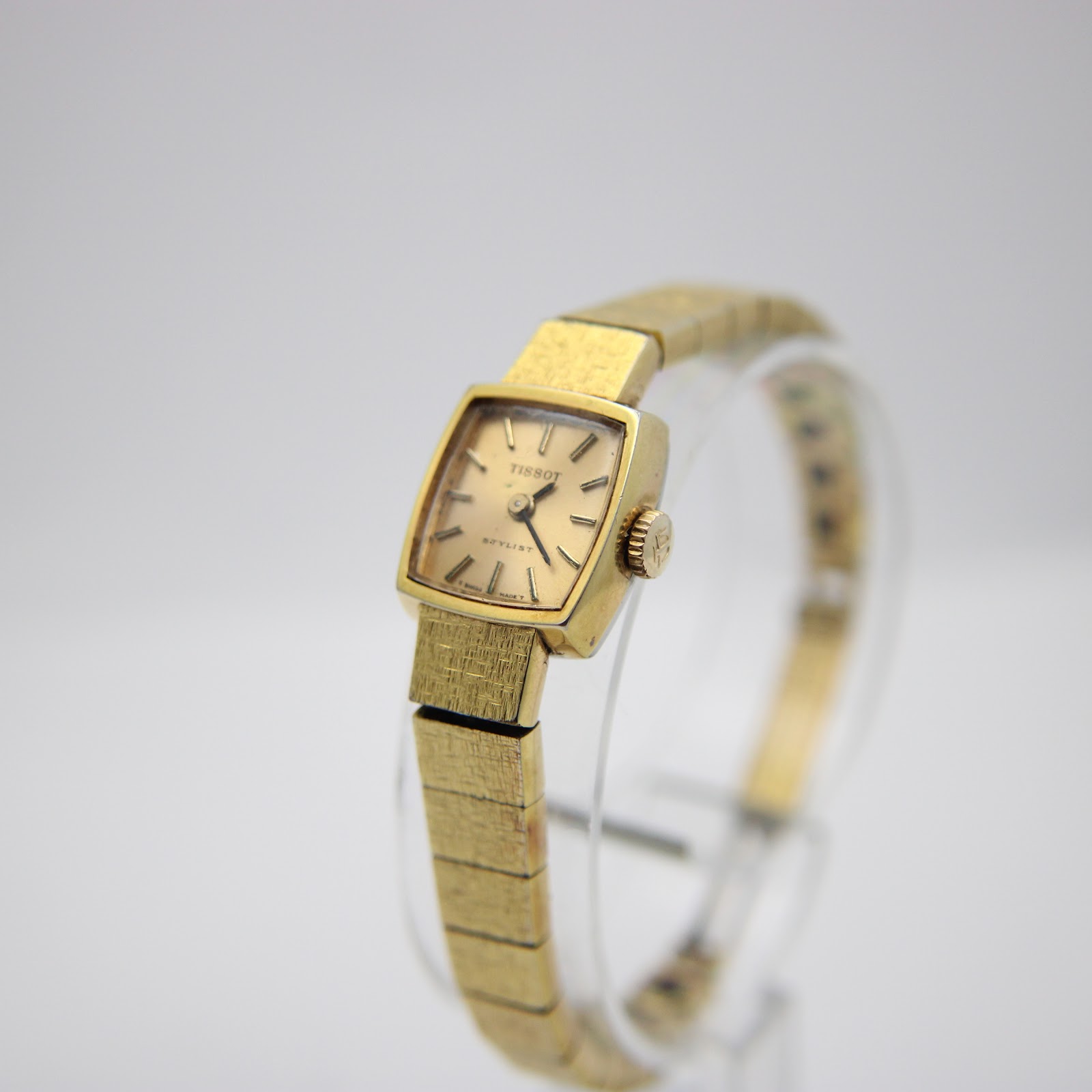 Angled front view of the Tissot Stylist on a clear display stand, showing the integrated gold-filled bracelet.