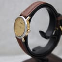 Left side view of the Tissot watch on a stand, showing the gold-tone crown and lugs, silver-tone case, and brown leather strap. The cream dial is visible.