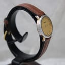 Right side view of the Tissot watch on a stand, showing the silver-tone case and gold-tone lugs, with the brown leather strap wrapped around. The dial is angled away.