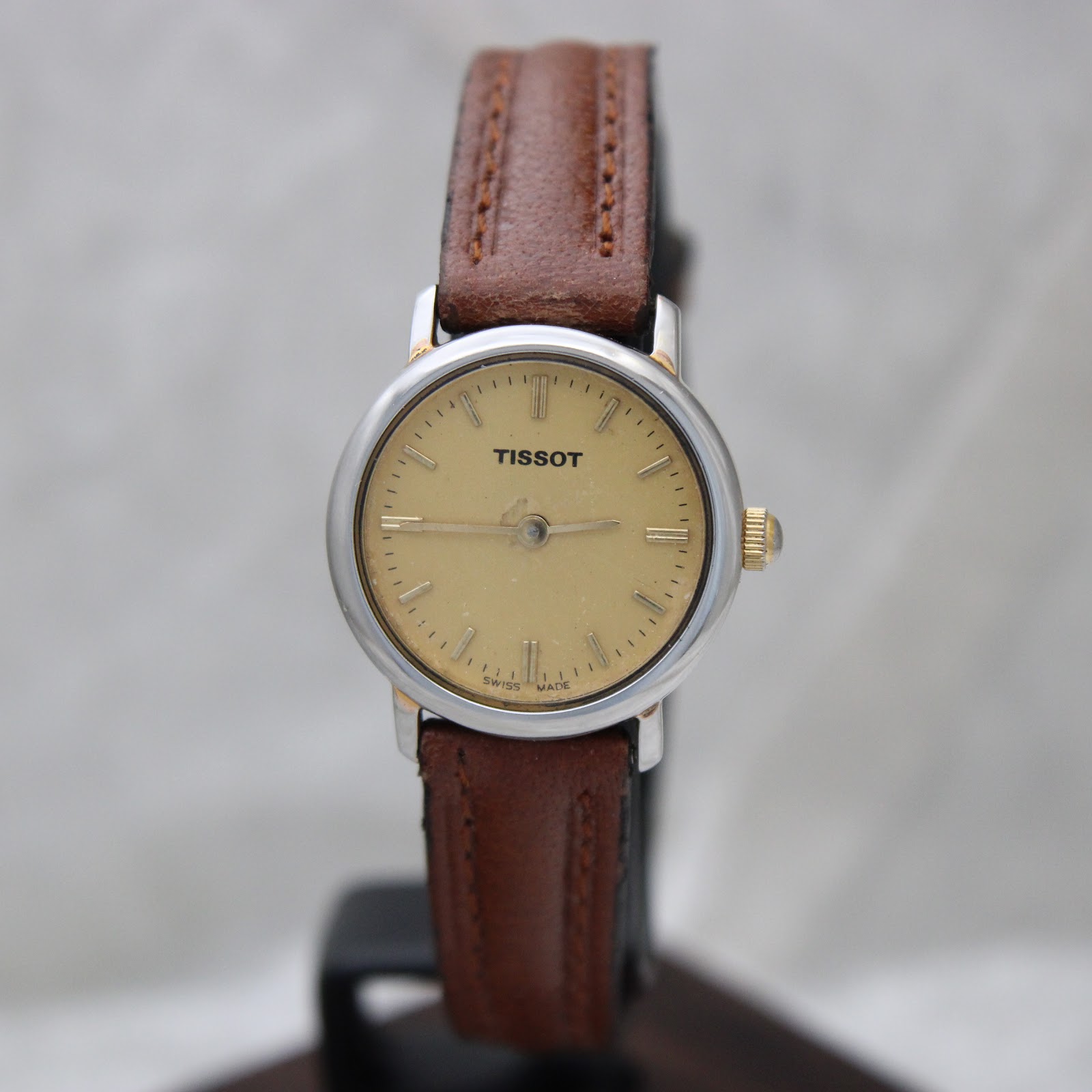 Front-facing view of the Tissot watch on a stand, clearly showing the warm cream dial with applied gold baton indices and "Tissot" logo, silver-tone bezel, and brown leather strap.