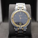 Front view of the Gucci 9000G watch on a grey display cushion, showing the black dial, gold-tone bezel, and two-tone bracelet.