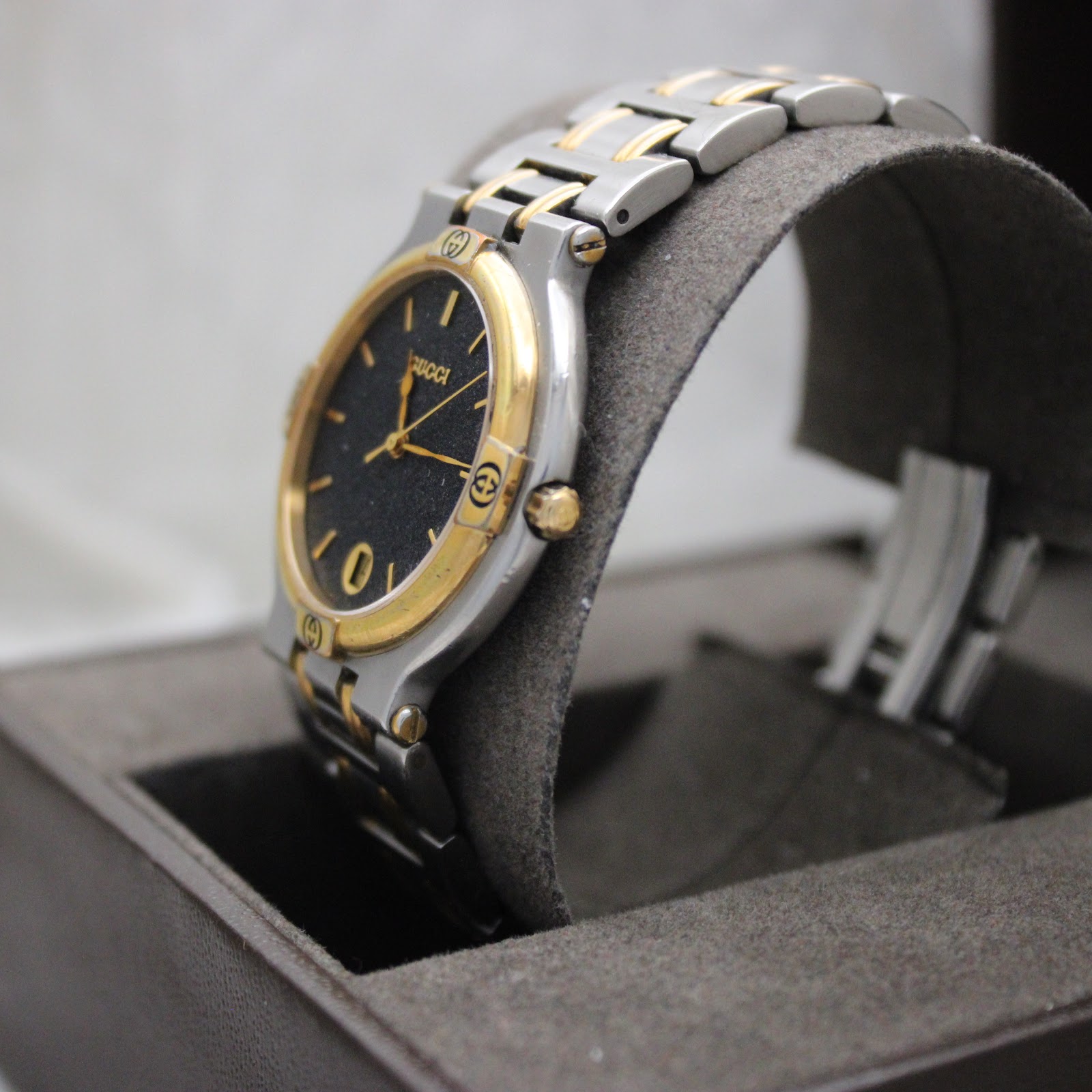 Angled side view of the Gucci 9000G watch on a grey display cushion, highlighting the case profile, gold crown, and two-tone bracelet.