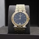 Front view of gold-tone Gucci 9200M watch with black dial and gold baton markers, displayed on a grey watch cushion inside a brown box.