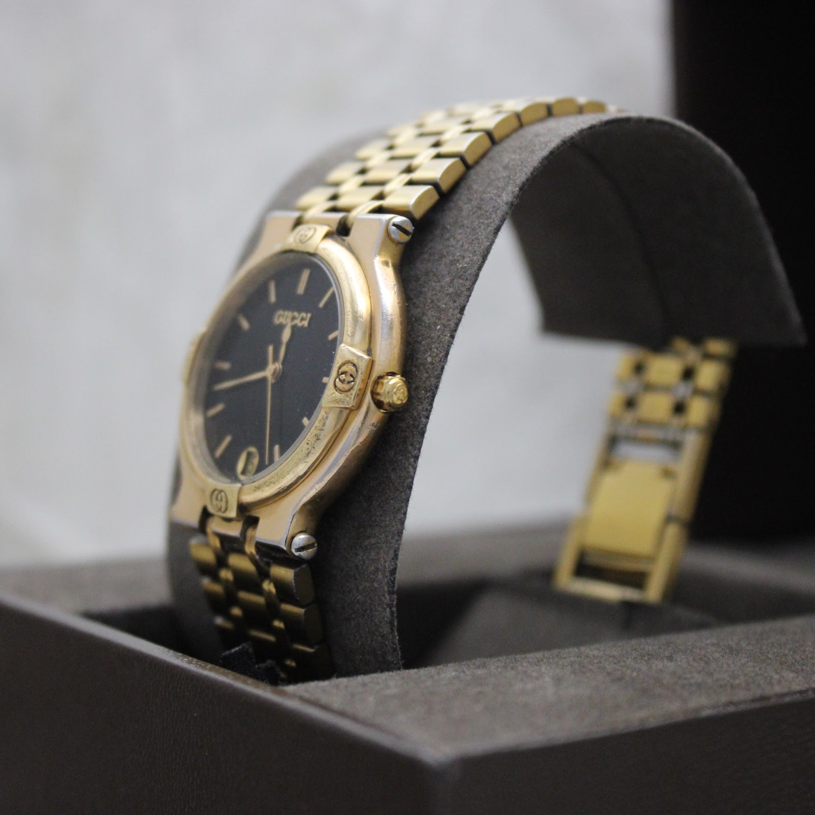 Angled side view of the gold-tone Gucci 9200M watch on a grey cushion, showing the crown and case profile.