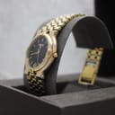 Angled side view of the gold-tone Gucci 9200M watch on a grey cushion, showing the crown and case profile.