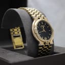 Another angled front view of the gold-tone Gucci 9200M watch on a grey cushion, highlighting the black dial and date window.