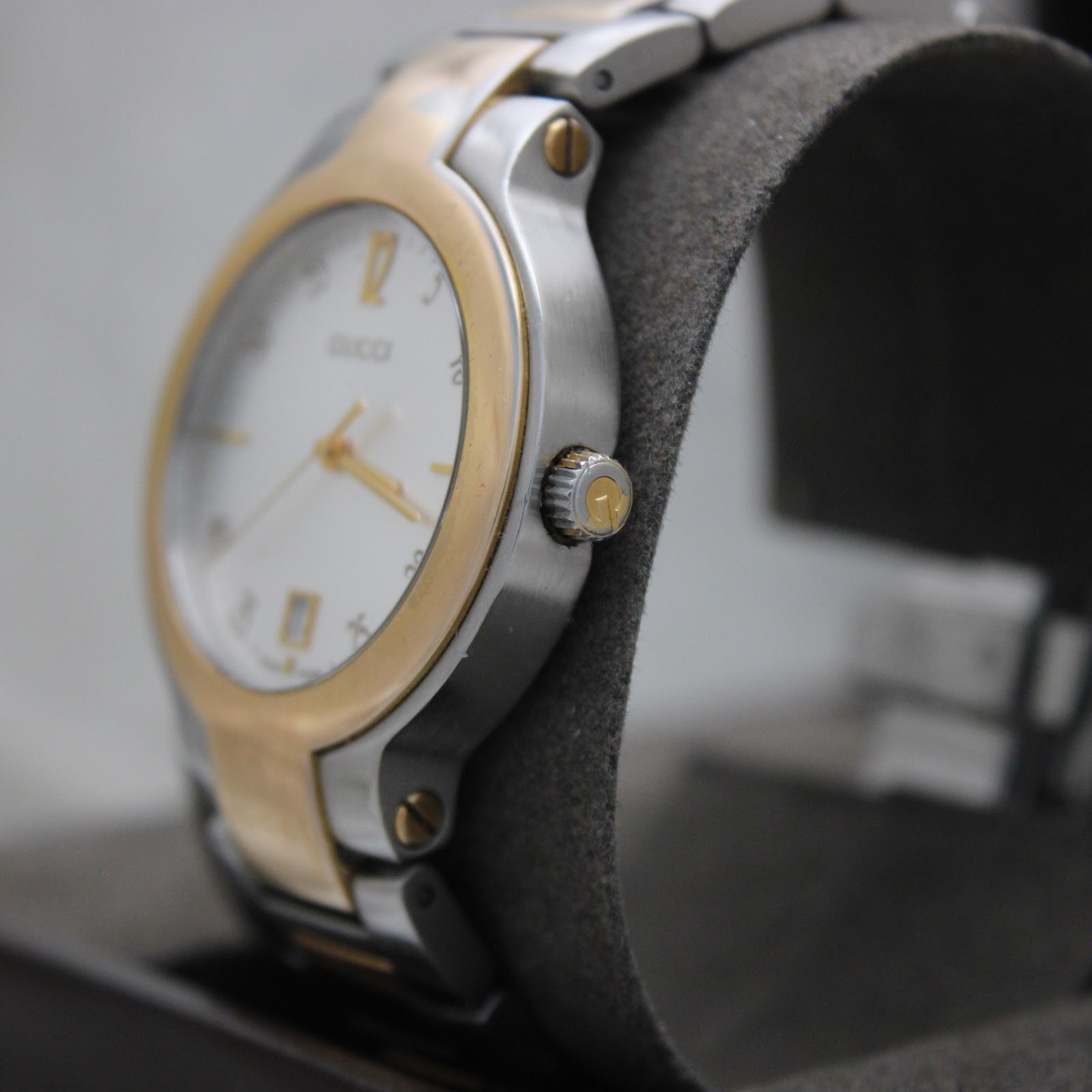 Angled side view of the Gucci 8900M watch, highlighting the gold-tone crown and the brushed stainless steel case on a grey display cushion.