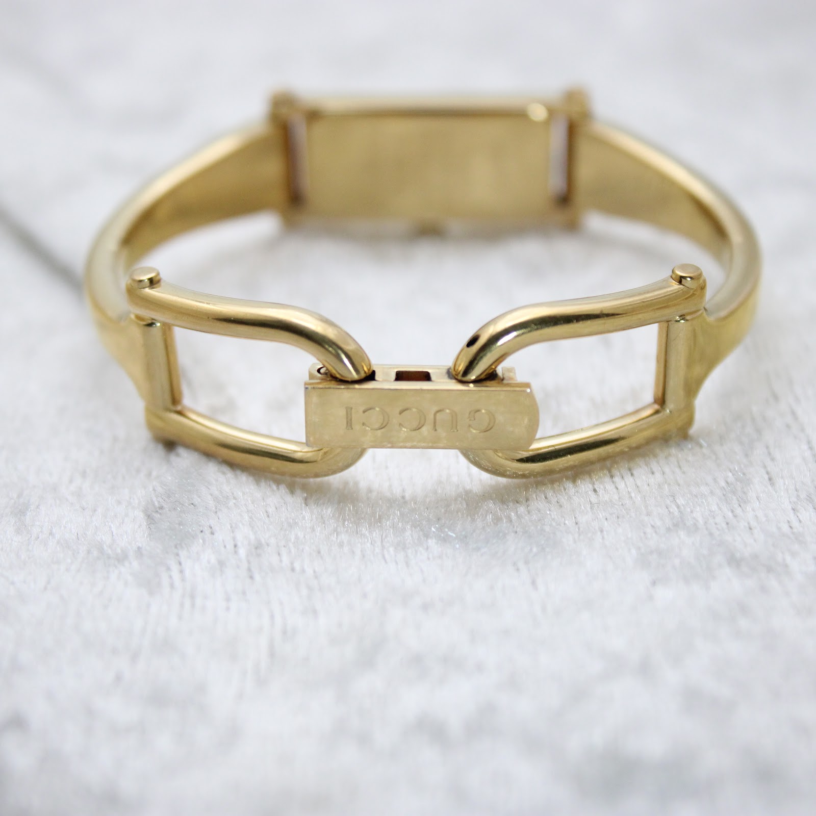 View of the open gold-tone bangle clasp, with "GUCCI" engraved on the fold-over component, resting on a white textured surface.