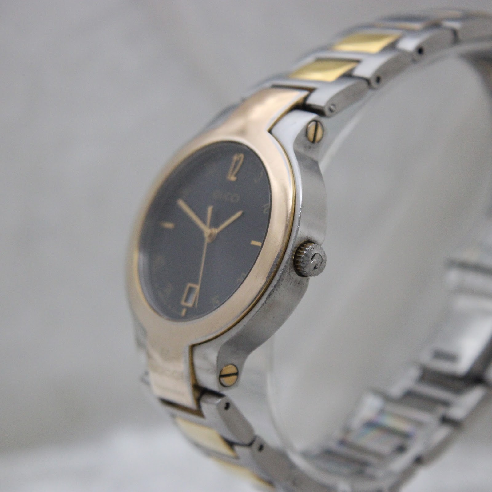 Angled side view of the Gucci 8900M watch, highlighting the crown with Gucci logo and the profile of the gold-tone bezel.