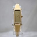 Front view of the gold-tone rectangular Gucci 1500L watch on a clear stand, showing the minimalist dial and gold bangle bracelet.