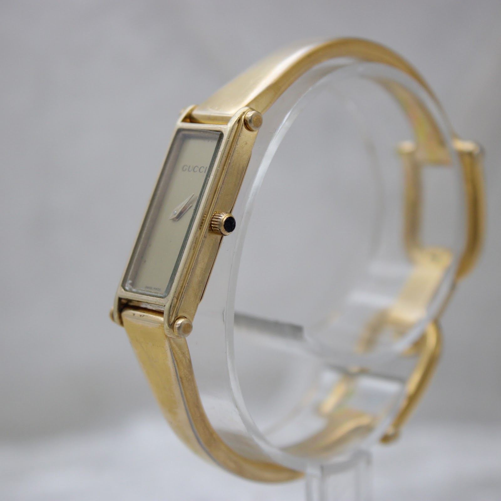 Angled view of the gold-tone Gucci 1500L watch, showing the side of the rectangular case, crown with blue cabochon, and bangle bracelet.