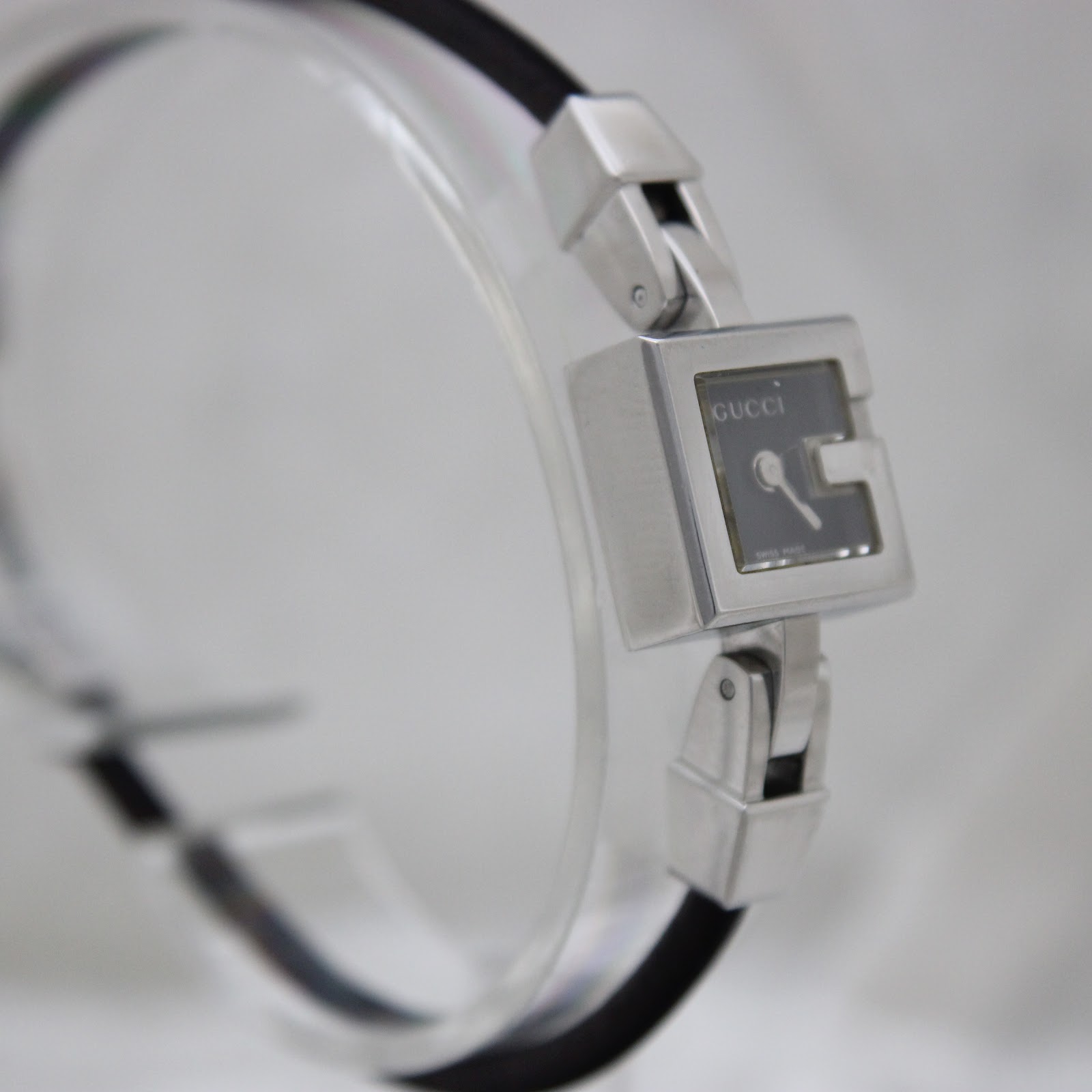 Side-angled view of the Gucci 102 watch on a clear stand, showing the minimalist black dial and the design of the stainless steel links.