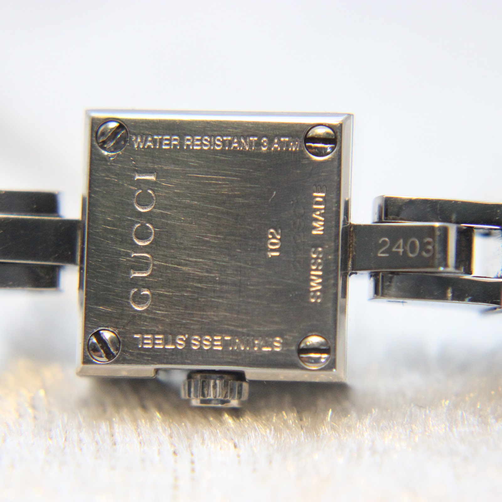 Close-up of the watch case back, revealing engravings: 'WATER RESISTANT 3 ATM', 'GUCCI', '102', 'SWISS MADE', 'STAINLESS STEEL', and model '2403'.