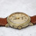 Close-up side view of the watch case and crown on a white surface, showing wear on the gold plating.
