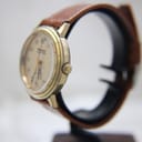 Side view of the watch on a display stand, showing the gold-plated crown and profile of the case.
