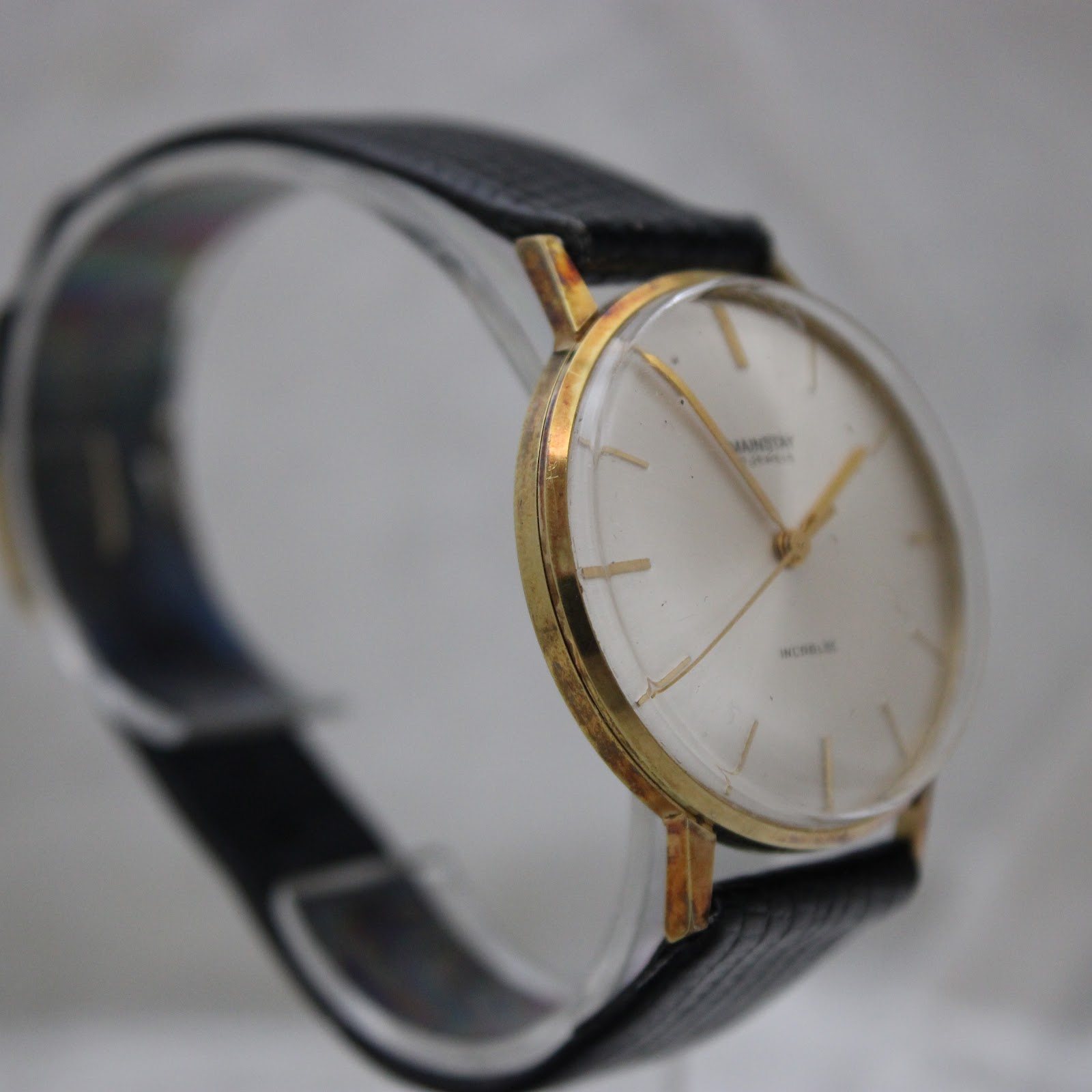 Three-quarter view of the Mainstay watch on a clear stand, highlighting the slim profile and black leather strap.