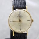 Front view of the gold-plated Mondaine watch with champagne sunburst dial, Roman numerals, and a black leather strap, on a watch stand.