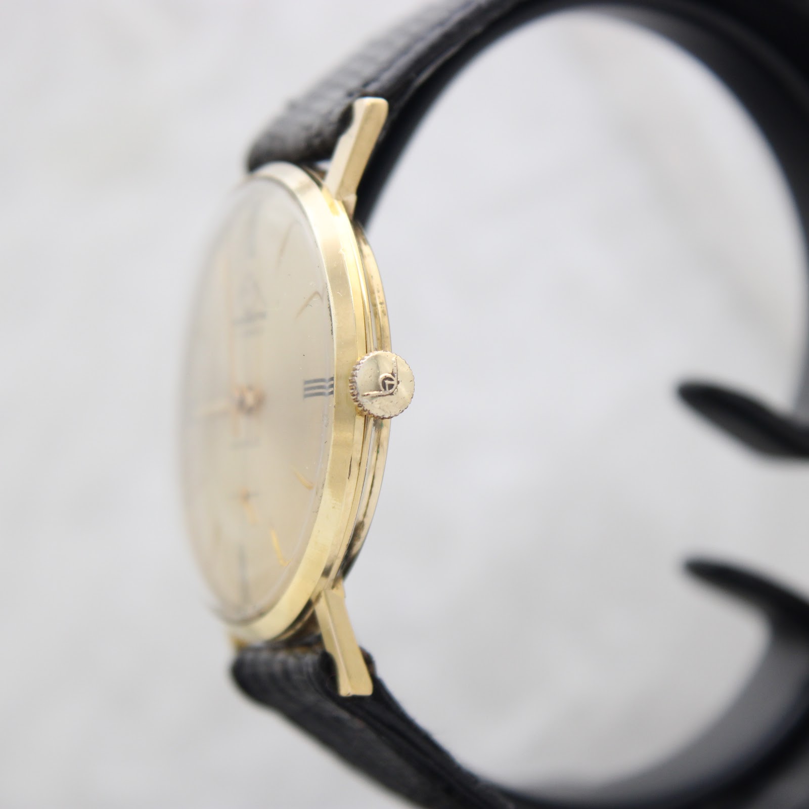 Side view of the Mondaine watch showing the slim gold-plated case and the signed crown, on a watch stand.