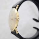 Side view of the Mondaine watch showing the slim gold-plated case and the signed crown, on a watch stand.