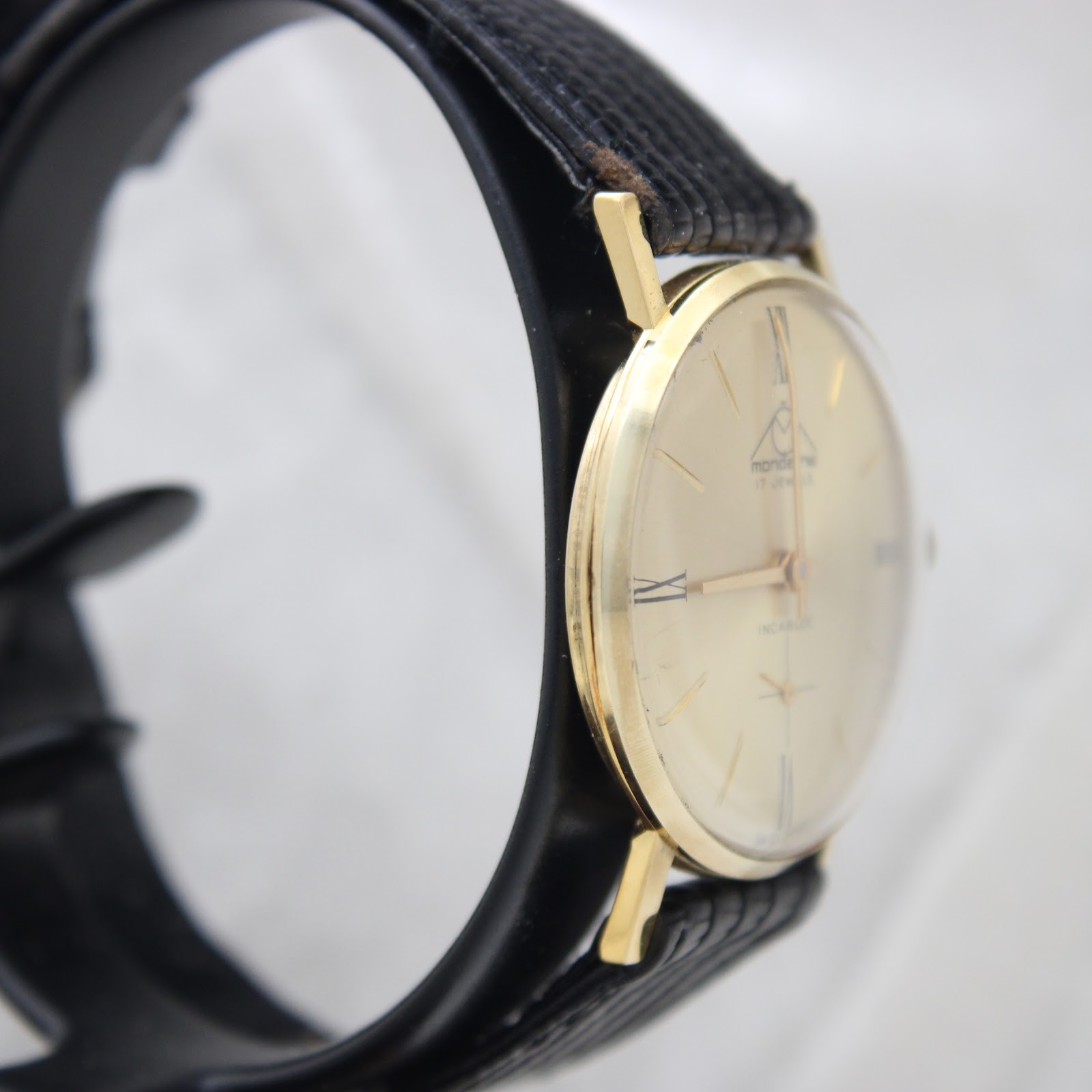 Angled side view of the Mondaine watch on a stand, highlighting the gold-plated case and the black leather strap.
