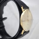 Angled side view of the Mondaine watch on a stand, highlighting the gold-plated case and the black leather strap.
