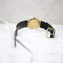 Back view of the Mondaine watch showing the plain gold-plated case back and the black leather strap with a gold buckle, on a white textured surface.