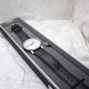 The Mondaine watch resting inside a simple, long grey presentation box, with its black leather strap laid out, suggesting careful storage.