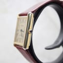 Side view of the Corteal watch showing the gold-tone case and cabochon crown, with the burgundy leather strap.