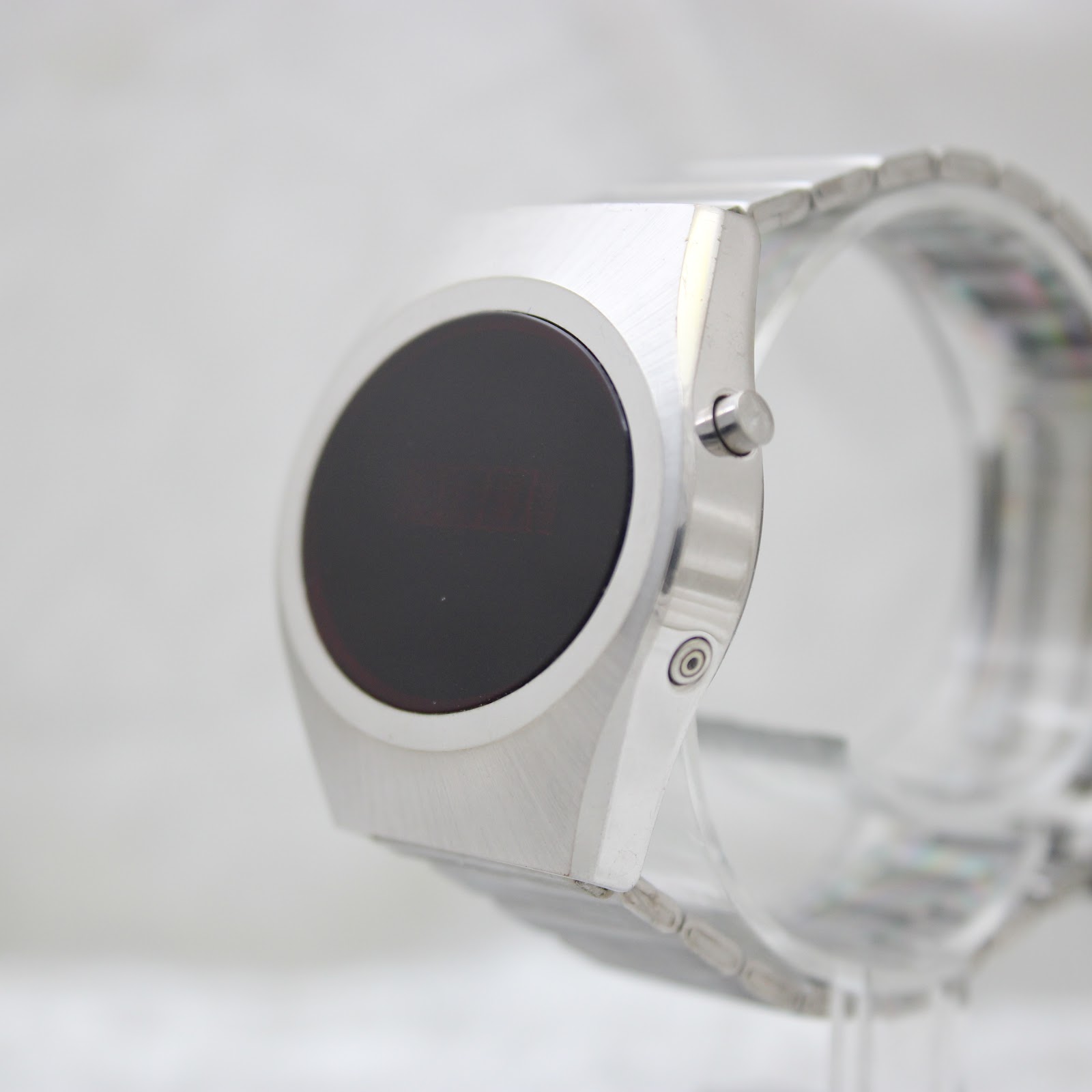Angled side view of the silver-tone LED digital watch on a clear stand, showing the push-button on the right side of the case.
