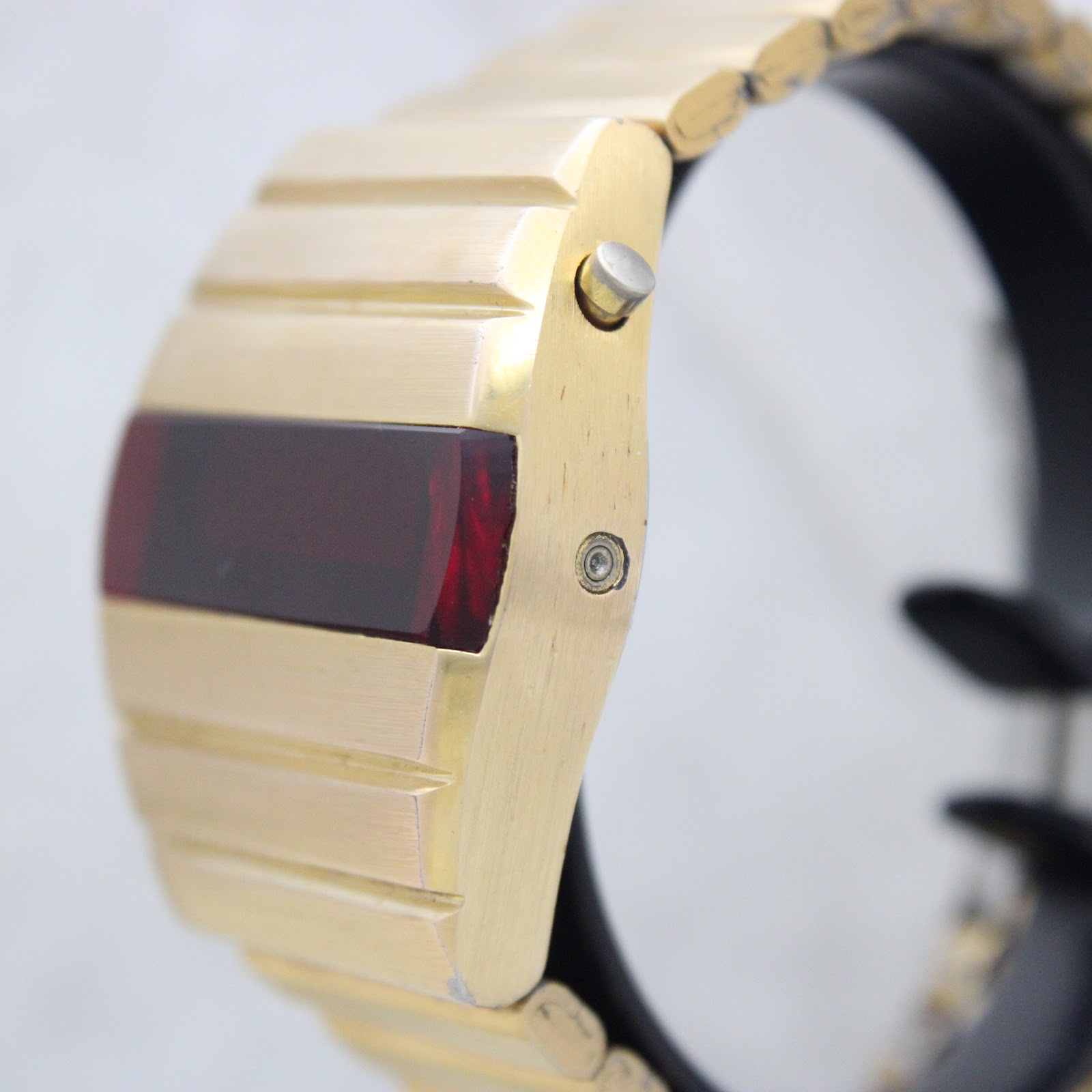 Side profile view of the watch case, showing the polished push-button crown for activating the display and a recessed setting pin, highlighting the gold plating.