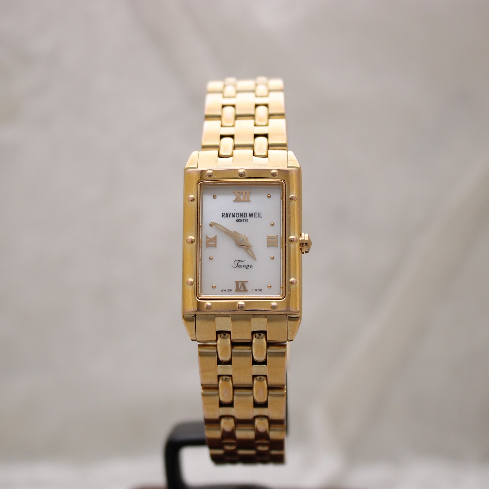 Straight-on front view of the Raymond Weil Tango watch with gold bracelet on a grey background