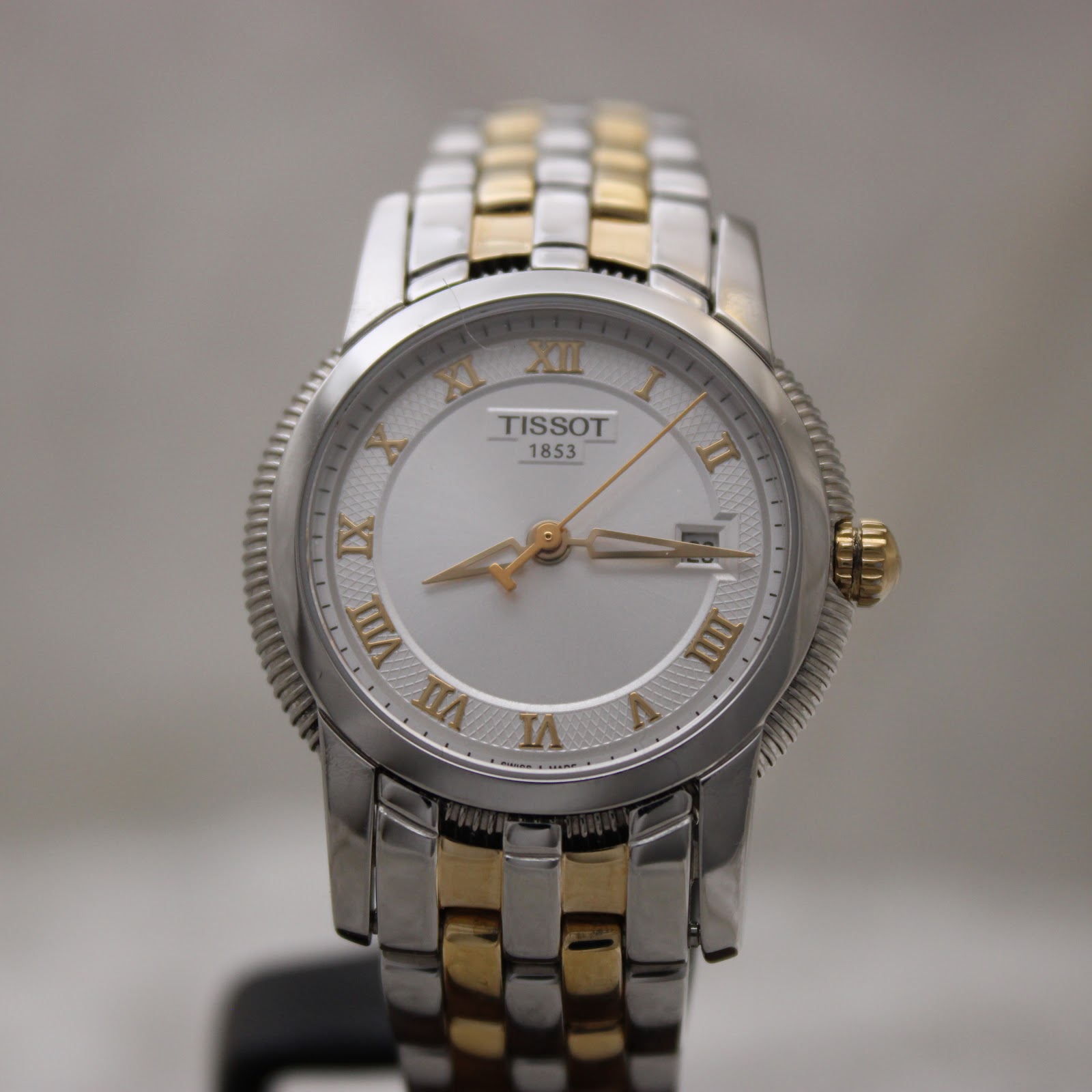 Close-up front view of the Tissot Ballade watch, highlighting the silver dial, gold Roman numerals, and date window.