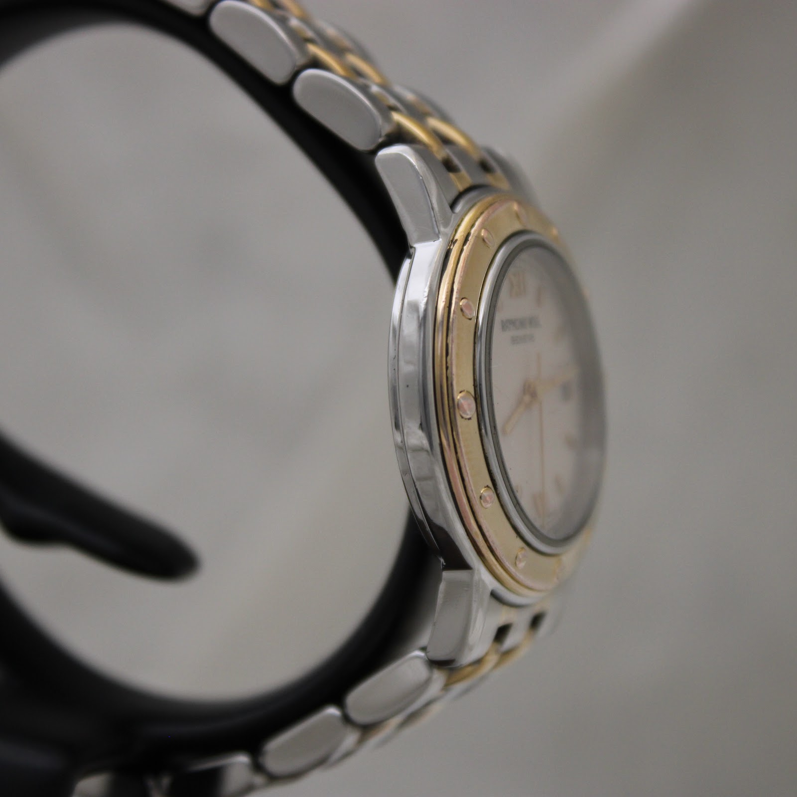 Angled side view of the Raymond Weil watch on a black stand, highlighting the gold-tone bezel with rivets and the brushed finish of the two-tone bracelet.