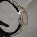 Angled side view of the Raymond Weil watch on a black stand, highlighting the gold-tone bezel with rivets and the brushed finish of the two-tone bracelet.
