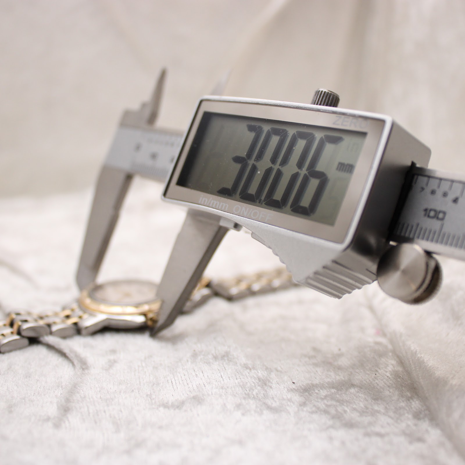 Digital caliper measuring the watch case, displaying a diameter of 30.06mm, on a light-coloured textured surface.
