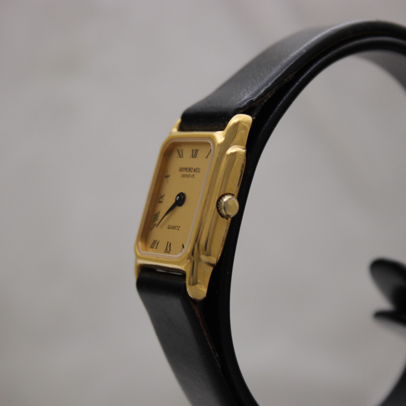 Angled view of the Raymond Weil 5700 watch, highlighting the side profile of the gold-tone case and crown, with the black leather strap visible.