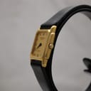Angled view of the Raymond Weil 5700 watch, highlighting the side profile of the gold-tone case and crown, with the black leather strap visible.