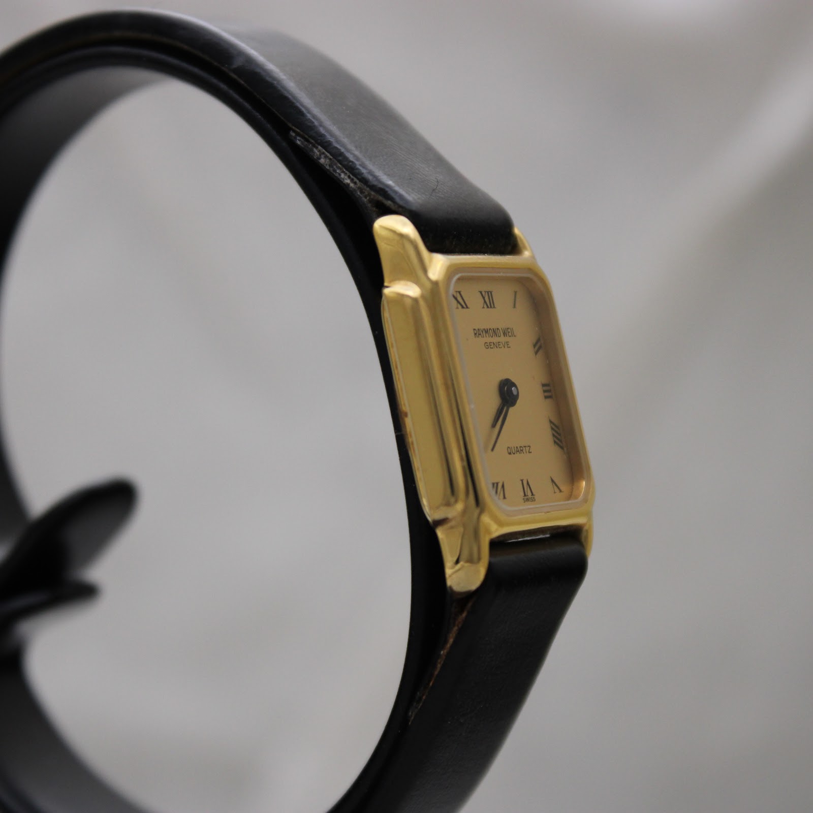 Another angled side view of the Raymond Weil Genève 5700 watch, focusing on the gold electroplated case and the minimalist design, set against a blurred background.