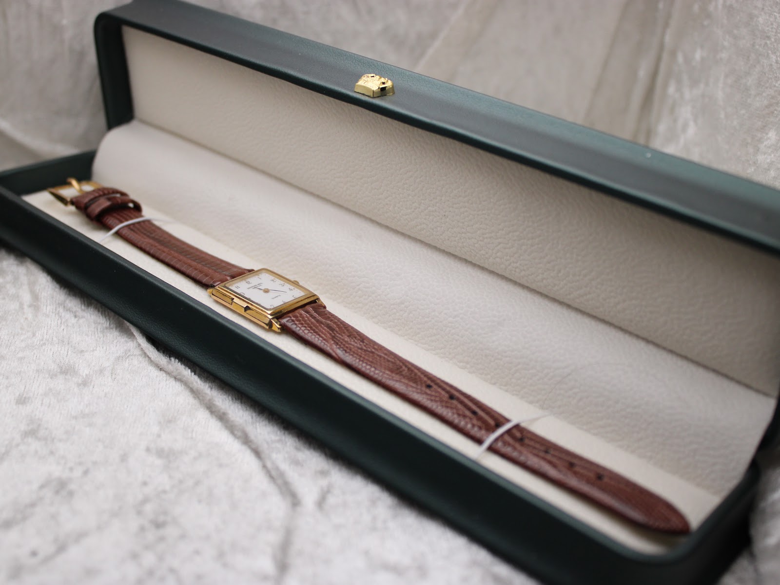 Overhead view of the Raymond Weil watch with its brown strap, presented inside an open, dark green watch box with a cream interior.