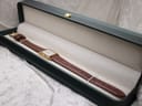 Overhead view of the Raymond Weil watch with its brown strap, presented inside an open, dark green watch box with a cream interior.