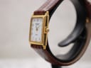 Angled side profile of the Raymond Weil watch on a display stand, showing the gold electroplated case, gold-tone crown, and brown leather strap.