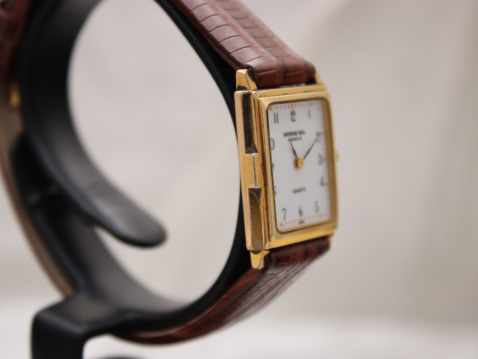 Opposite angled side profile of the Raymond Weil watch on a display stand, highlighting the rectangular gold case and textured brown strap.